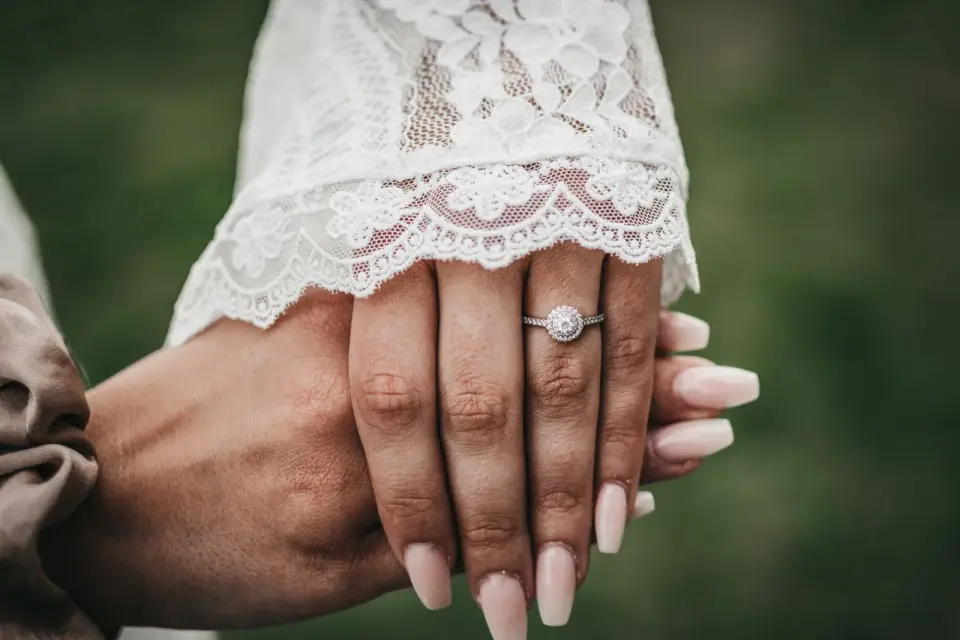 Bride’s hand with a diamond halo engagement ring and a lace sleeve, held by the groom