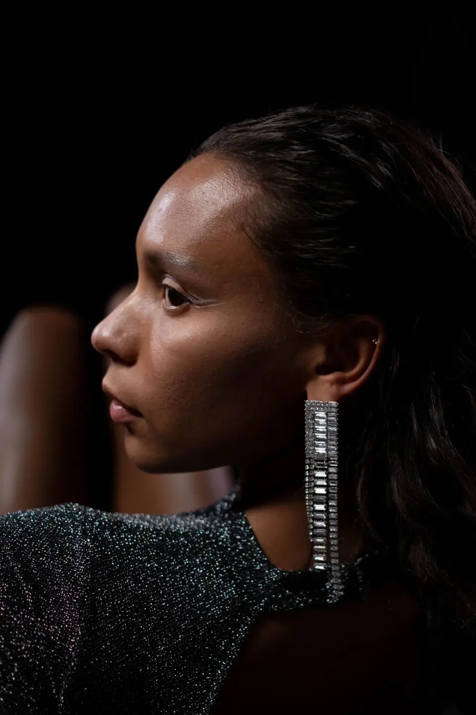 Woman in profile wearing a long baguette-crystal statement earring on a dark background