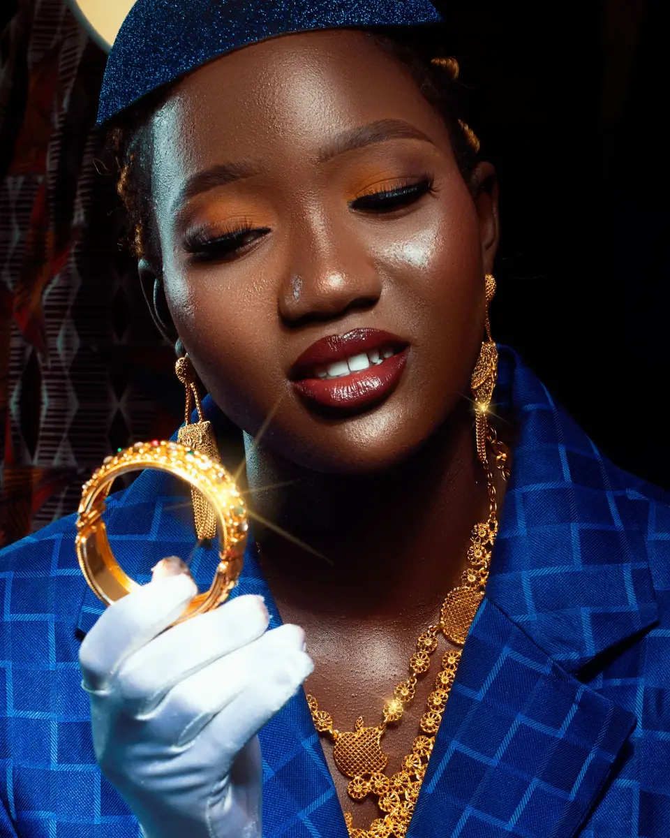 Woman in a blue blazer holding up a gold bangle in a white glove, gold necklace and drop earrings