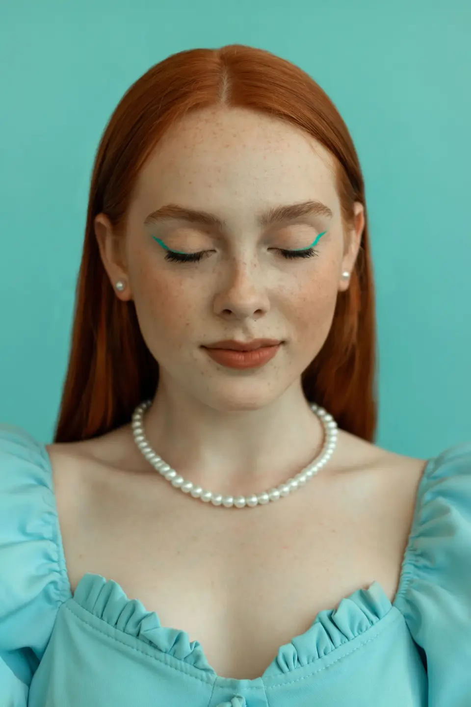 Red-haired woman wearing a pearl necklace and pearl stud earrings against a turquoise wall