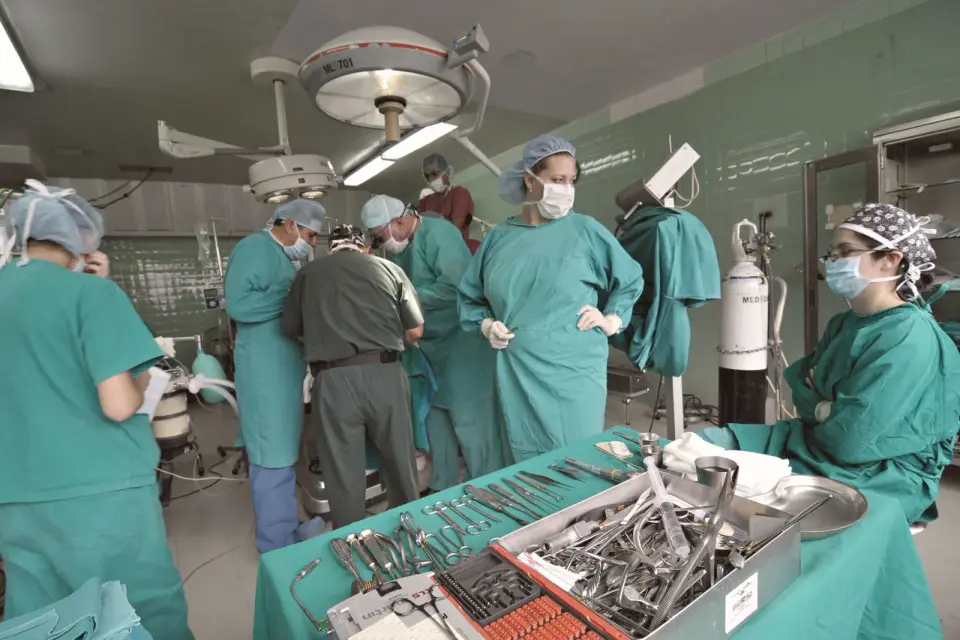 Surgical team in green scrubs, caps and masks around an operating table, with a tray of steel instruments in the foreground