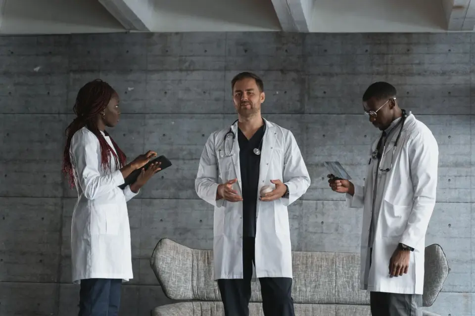 Three doctors in white coats talking, two holding tablets, against a concrete wall