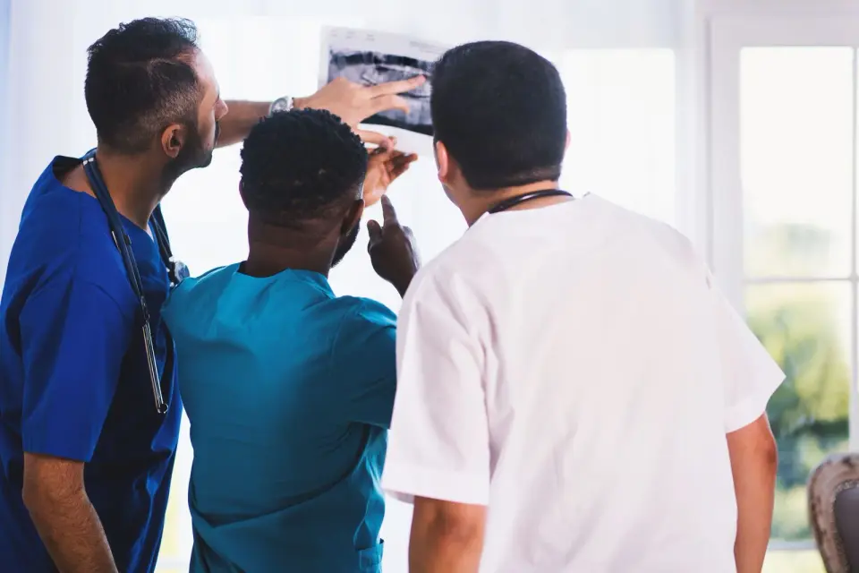 Three clinicians in scrubs and a white coat reviewing an X-ray film held up to the light