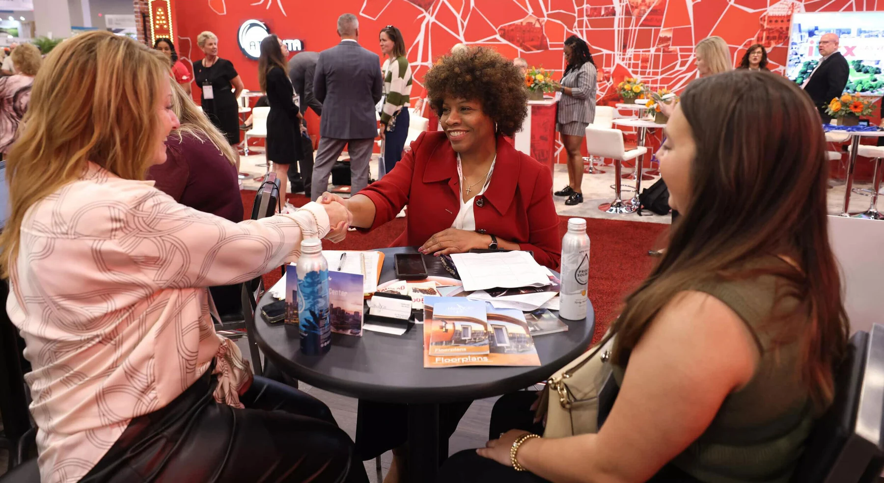Two women shake hands across a booth meeting table at IMEX America.