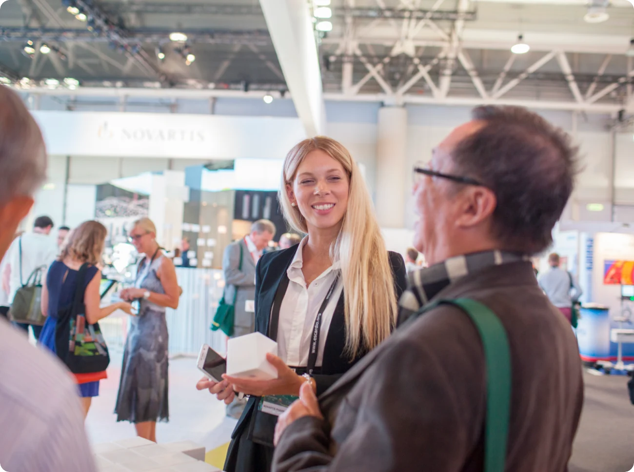 A smiling woman exhibitor in a black blazer welcomes visitors at a busy trade show stand.