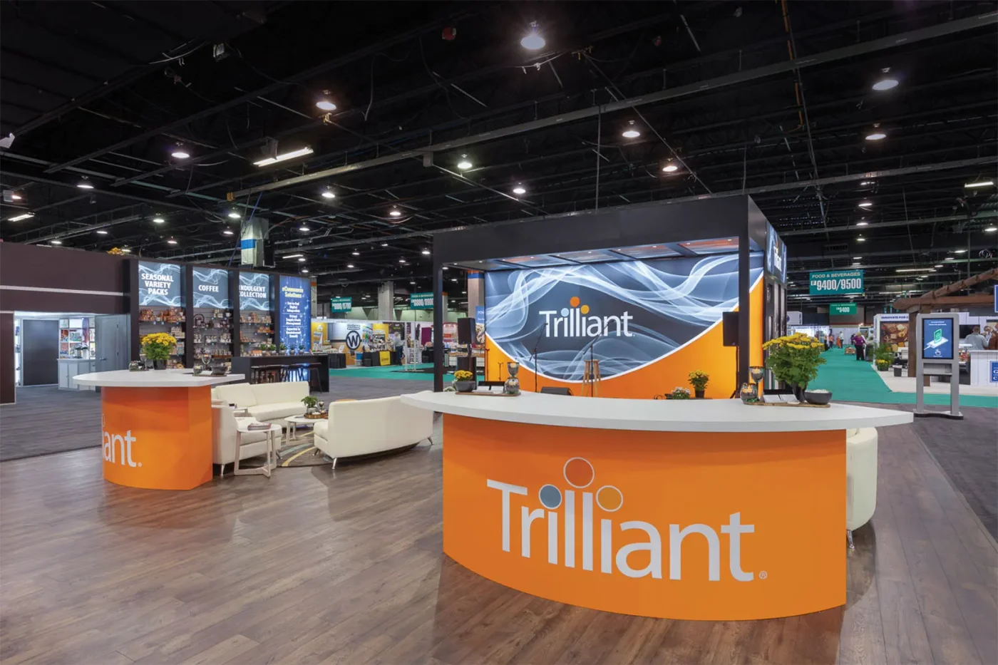 Trilliant exhibit by Purple Exhibits Trilliant exhibit from the Purple Exhibits portfolio, with curved orange welcome counters, an illuminated brand wall, and a lounge meeting area.