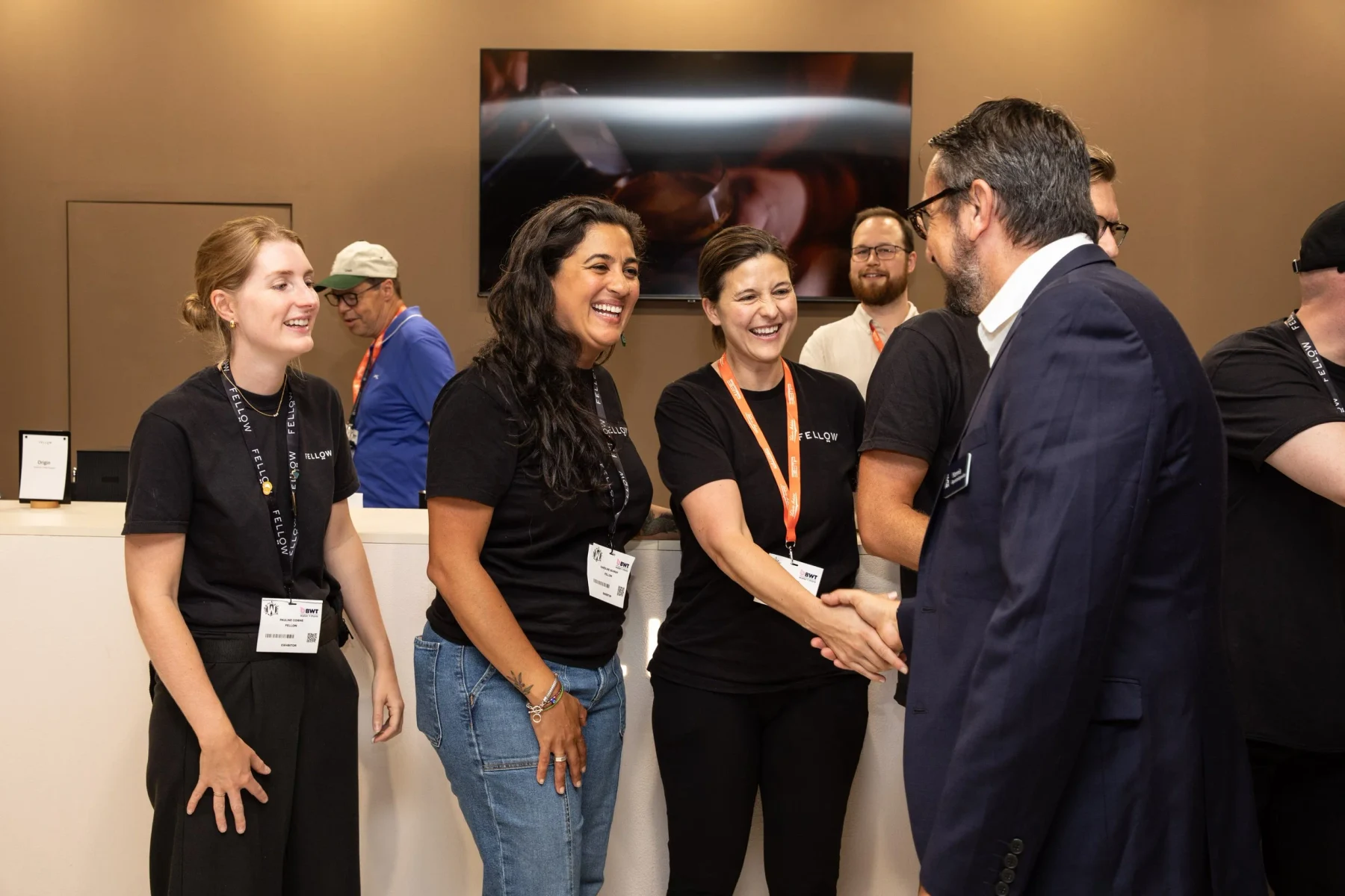 The Fellow booth team welcomes a World of Coffee visitor Three women on the Fellow booth team welcome a visitor with a handshake at World of Coffee Geneva.