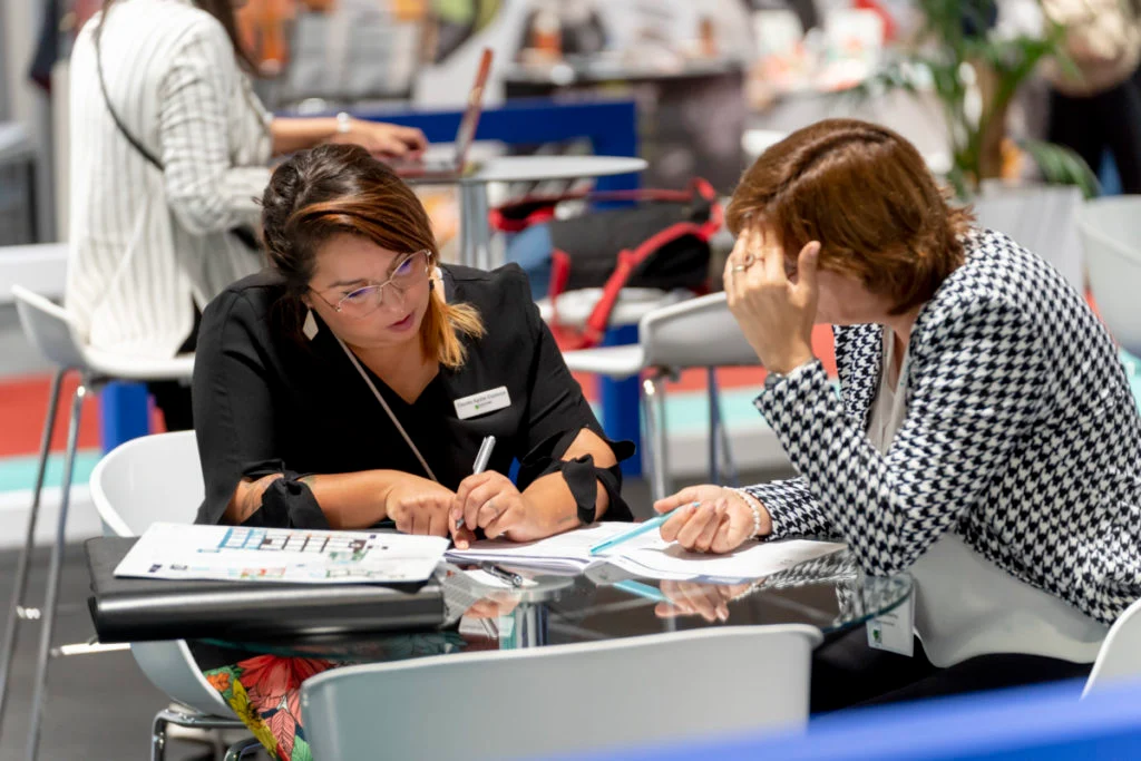 Reviewing plans together on the exhibition floor Two women review printed plans and write notes together at a table on the exhibition floor.