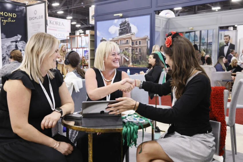 A meeting-table handshake at IMEX America Women shake hands across a booth meeting table with a tablet at IMEX America.