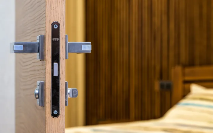 A modern wooden door with a metal handle and lock opens toward a bedroom.