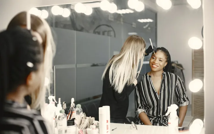 A makeup artist applies cosmetics to a smiling woman at an illuminated mirror.