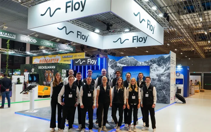 Floy team posing in a medical-technology exhibit beneath large suspended white signs, with imaging workstations and mountain graphics behind them.