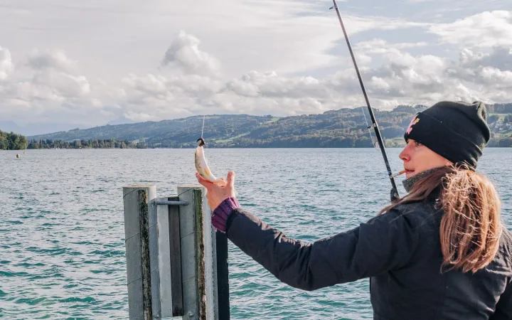 A woman fishes beside a lake with mountains in the distance.
