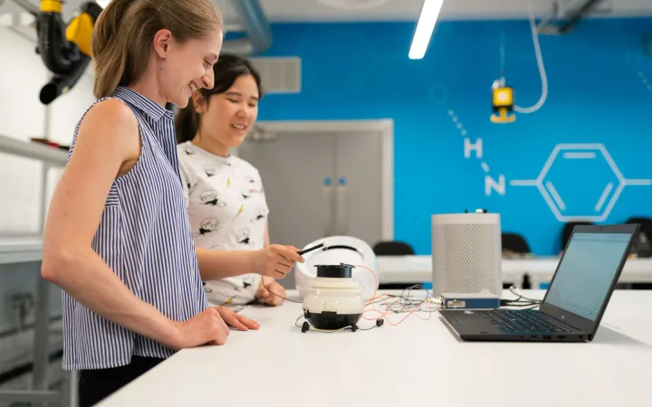 Two women investigate an electronic prototype and laptop at a bright laboratory bench.