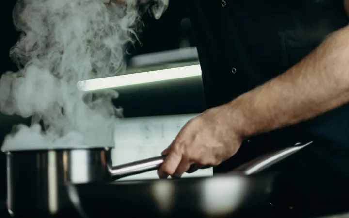 A chef tastes a dish in a professional kitchen with steam rising.
