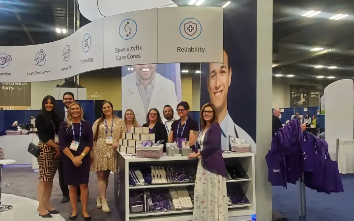 People posing at SpecialtyRx's exhibit around a giveaway counter, beneath a curved canopy listing pharmacy-service commitments.