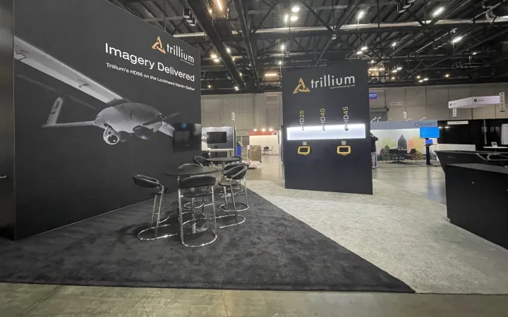 Trillium Engineering exhibit with dark aircraft graphics, illuminated camera-sensor displays, a reception counter and high meeting tables.