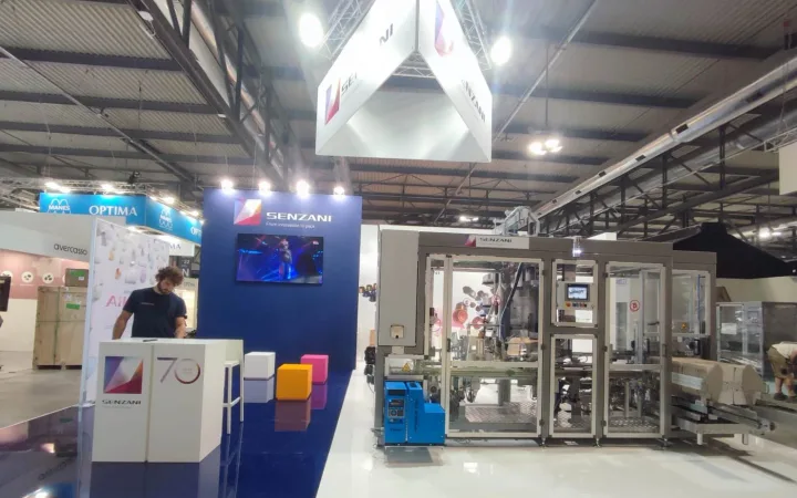 Senzani exhibit with a large enclosed packaging machine, a control screen, purple branded wall, suspended cube sign and a person at the reception counter.