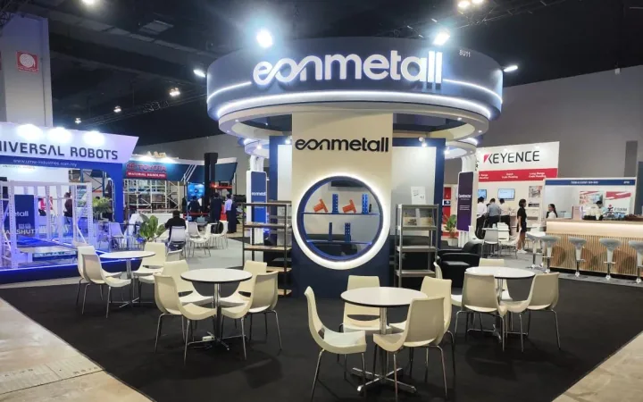 Eonmetall exhibit with an illuminated circular overhead sign, a central branded display, round meeting tables and white chairs.
