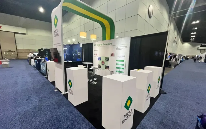 Mitsui Kinzoku exhibit with a green-and-yellow overhead arch, white display plinths and a wall about copper powder for additive manufacturing.