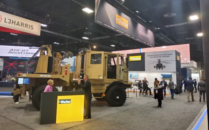 Parker defense exhibit with a military vehicle, large suspended brand signs, illuminated reception counter and visitors beside technical product displays.