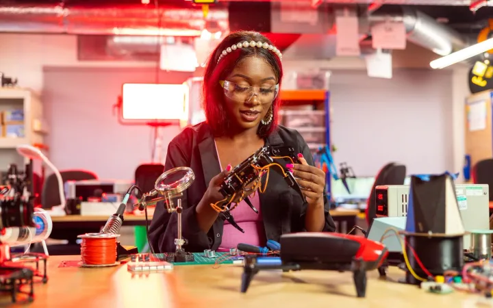 A woman works with a small drone and electronic components at a workbench.