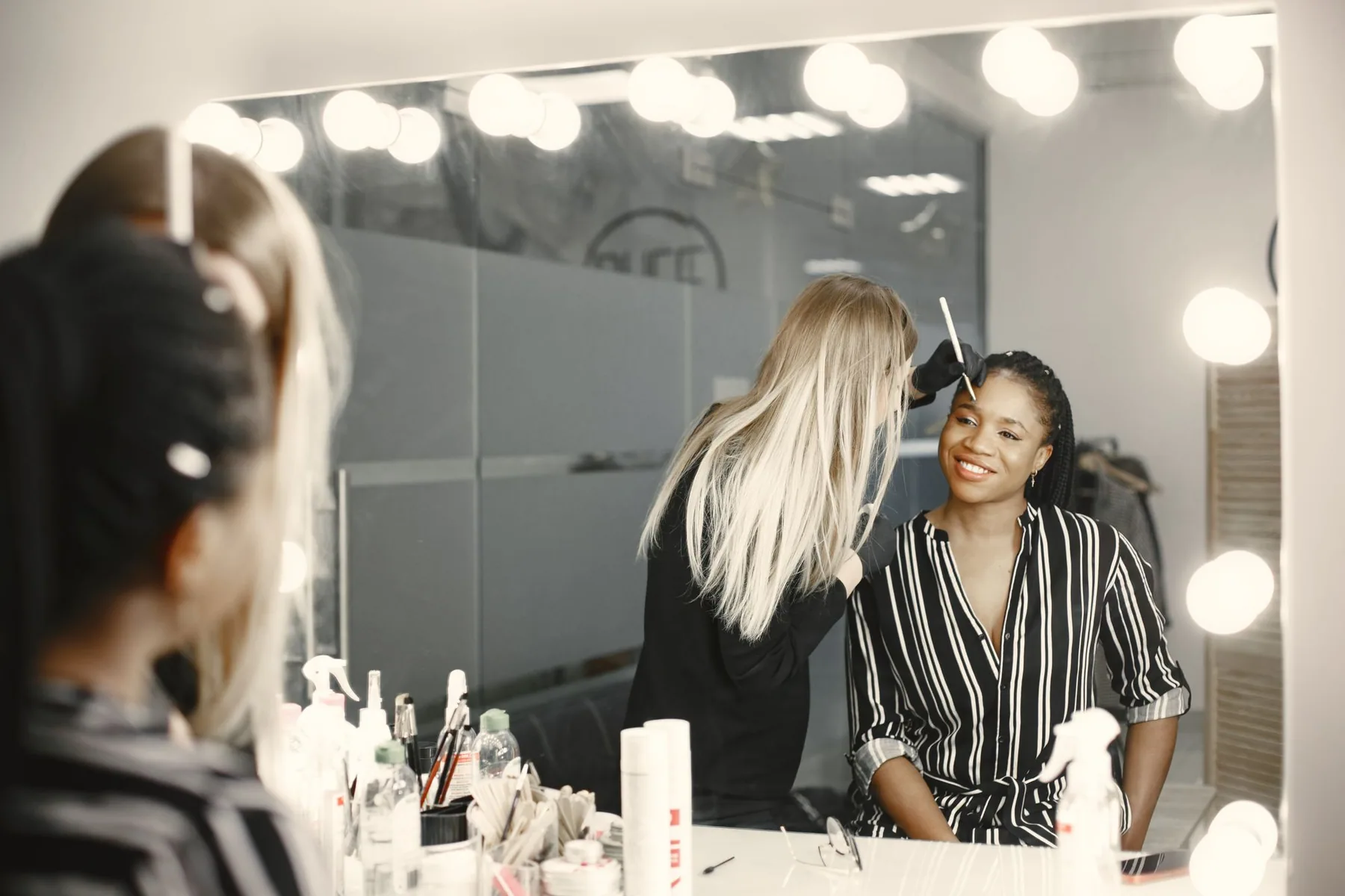 A makeup artist applies cosmetics to a smiling woman at an illuminated mirror.