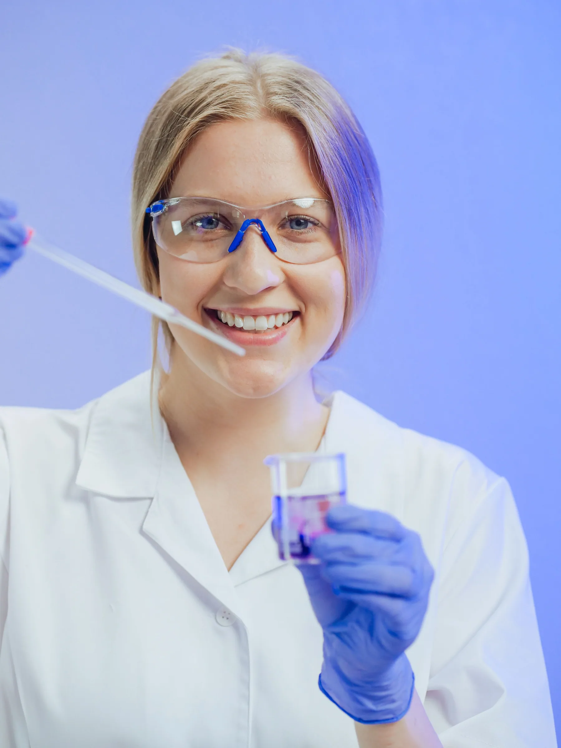 A smiling woman scientist in a lab coat holds laboratory glassware and a pipette.