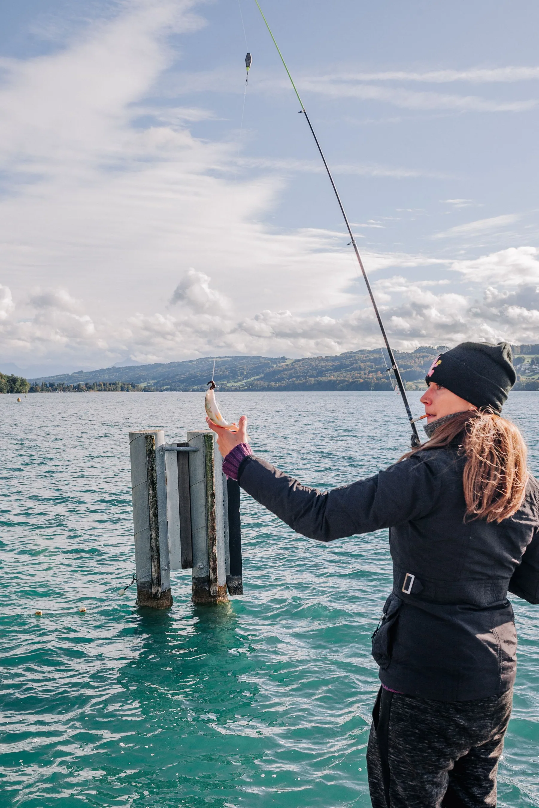 A woman fishes beside a lake with mountains in the distance.