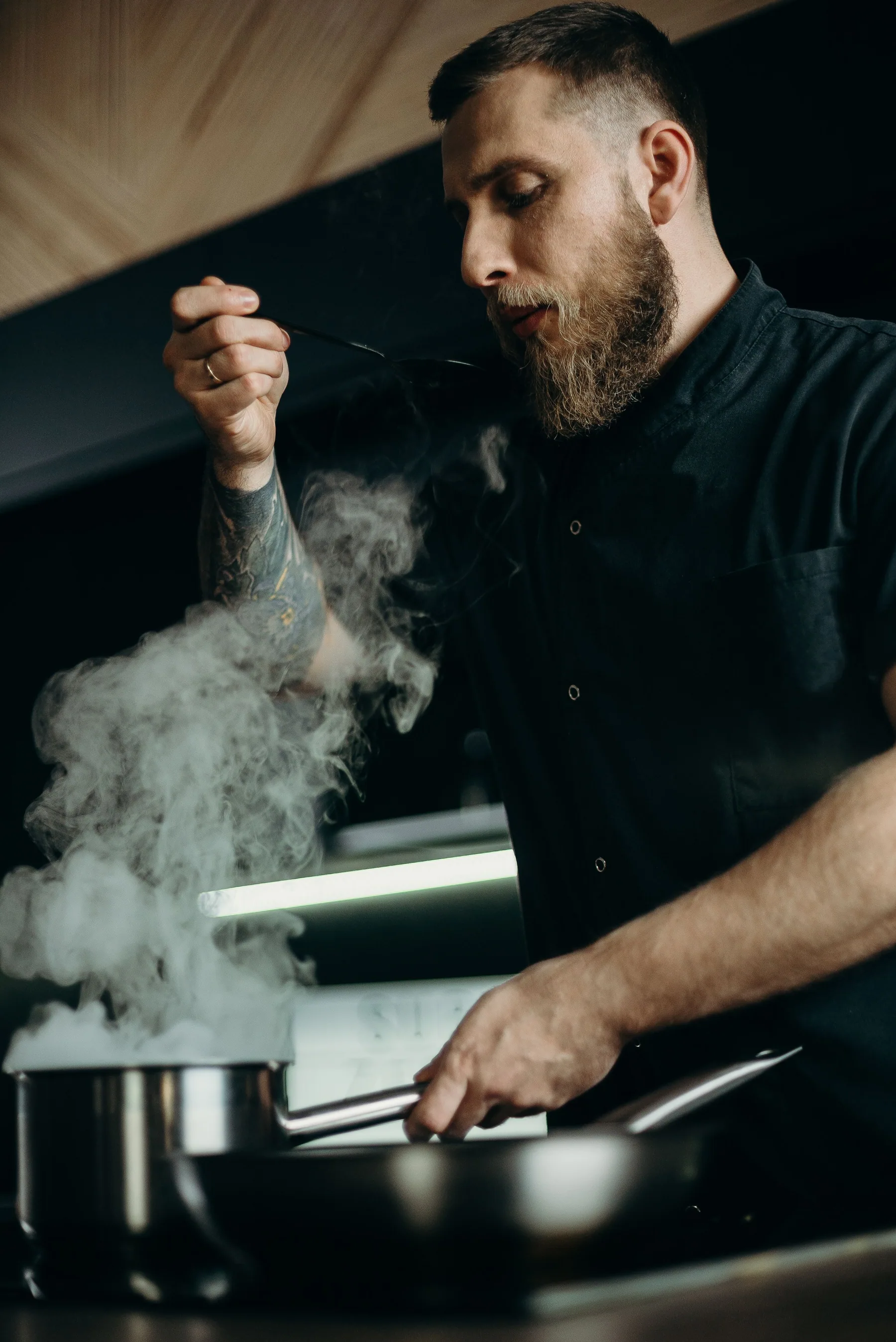 A chef tastes a dish in a professional kitchen with steam rising.
