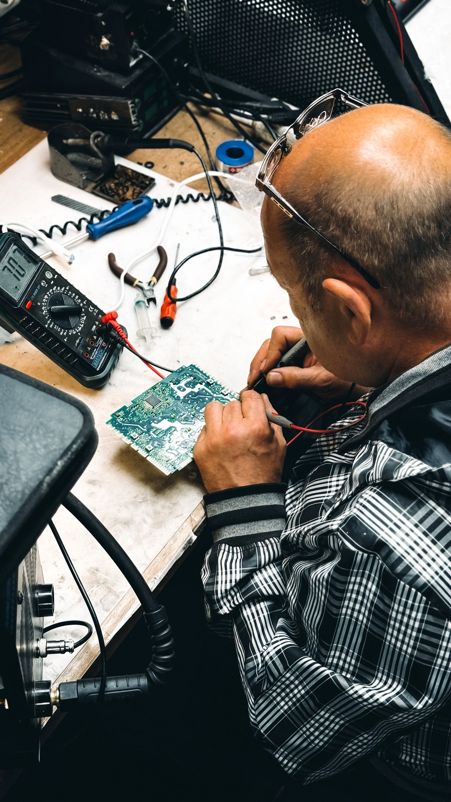 A technician examines a circuit board at an electronics workbench.