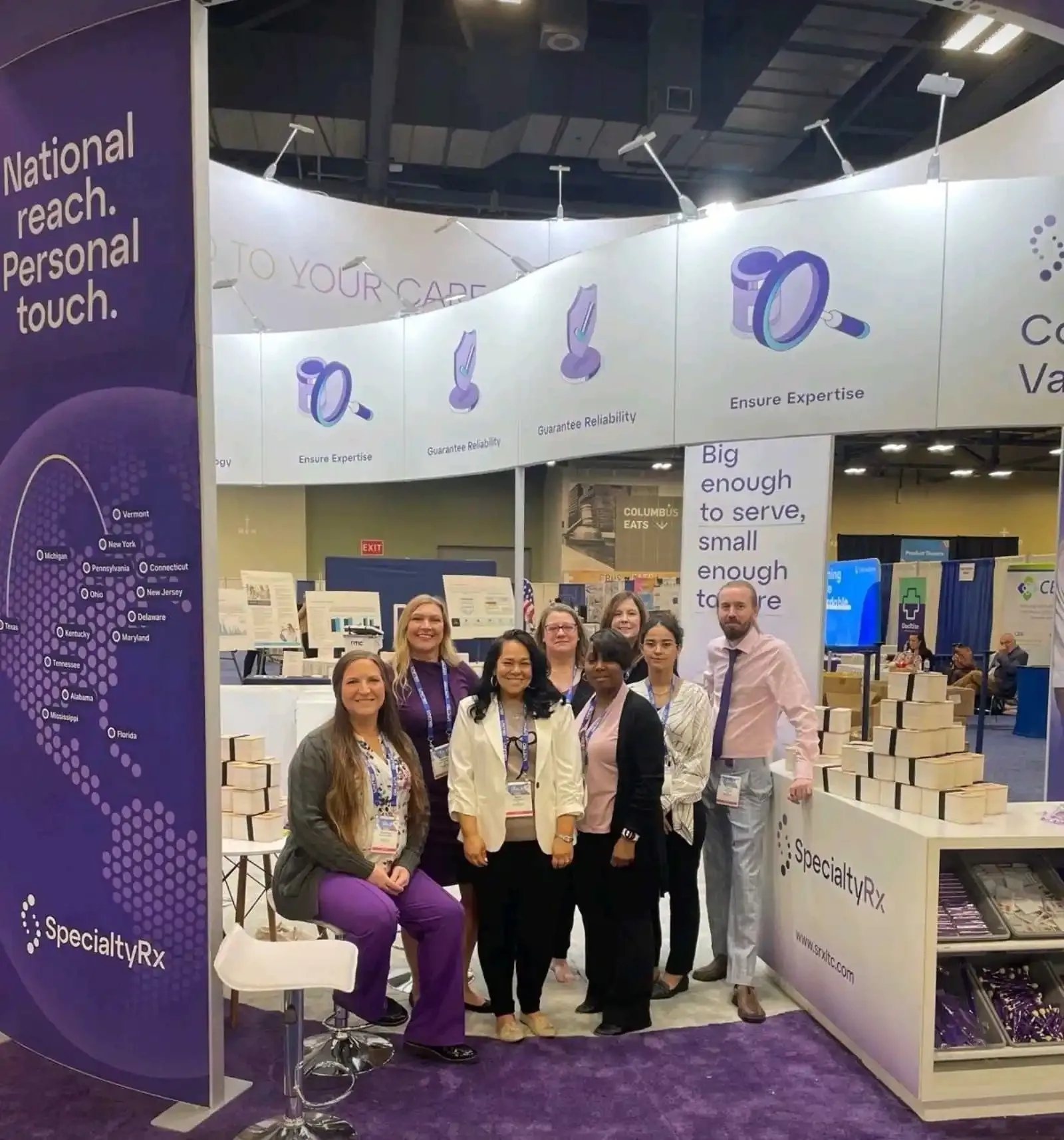 SpecialtyRx staff at a counter within a purple and white pharmacy-services exhibit with overhead branding.