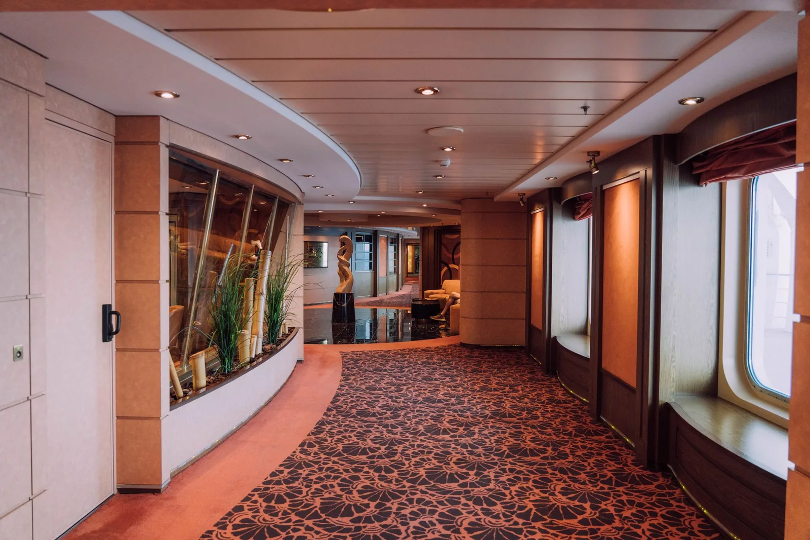 Cruise-ship corridor with patterned carpet, warm wall panels, recessed lighting and windows along one side.