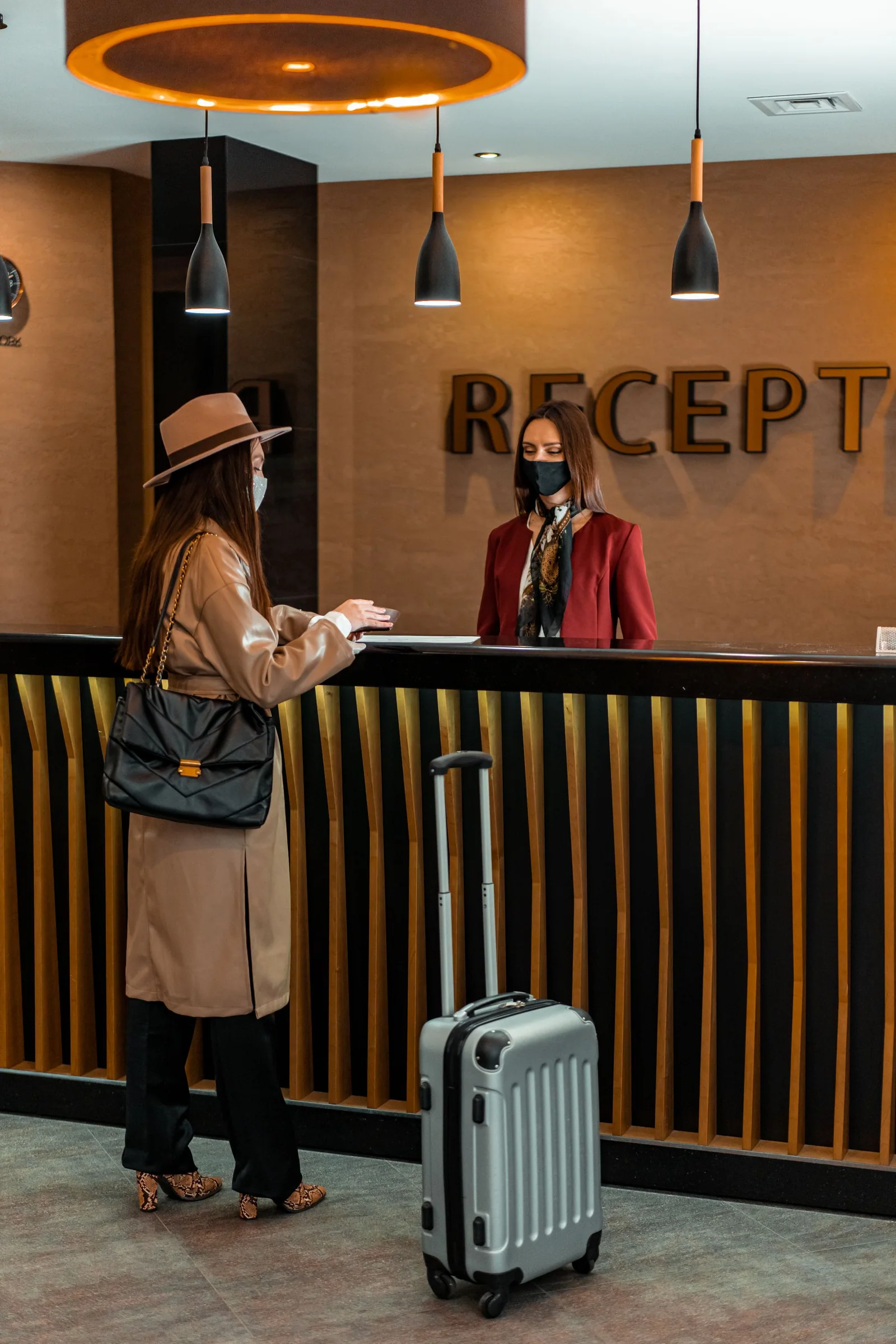 A hotel guest in a tan coat and hat speaks with a receptionist in a red blazer; both wear face masks.