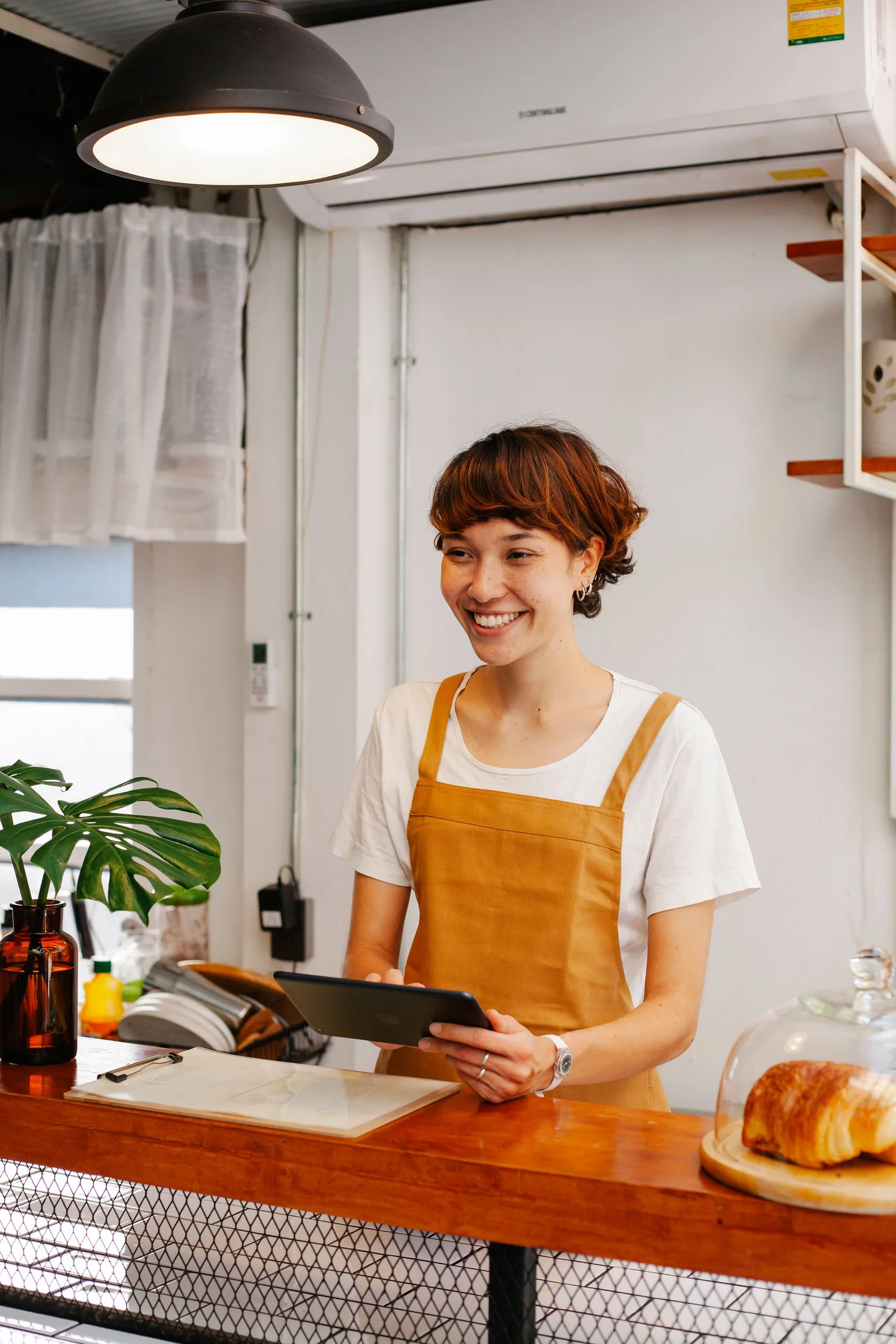 Smiling adult bakery worker in an apron holding a tablet beside a pastry display.