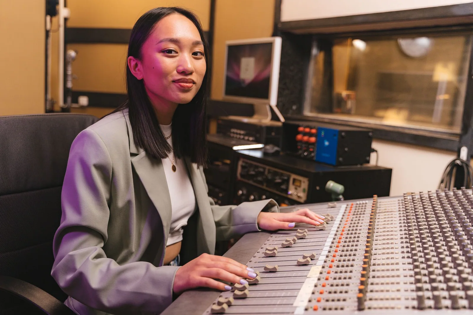 A woman in a light blazer sits at a studio audio mixing console with her hands on the faders.