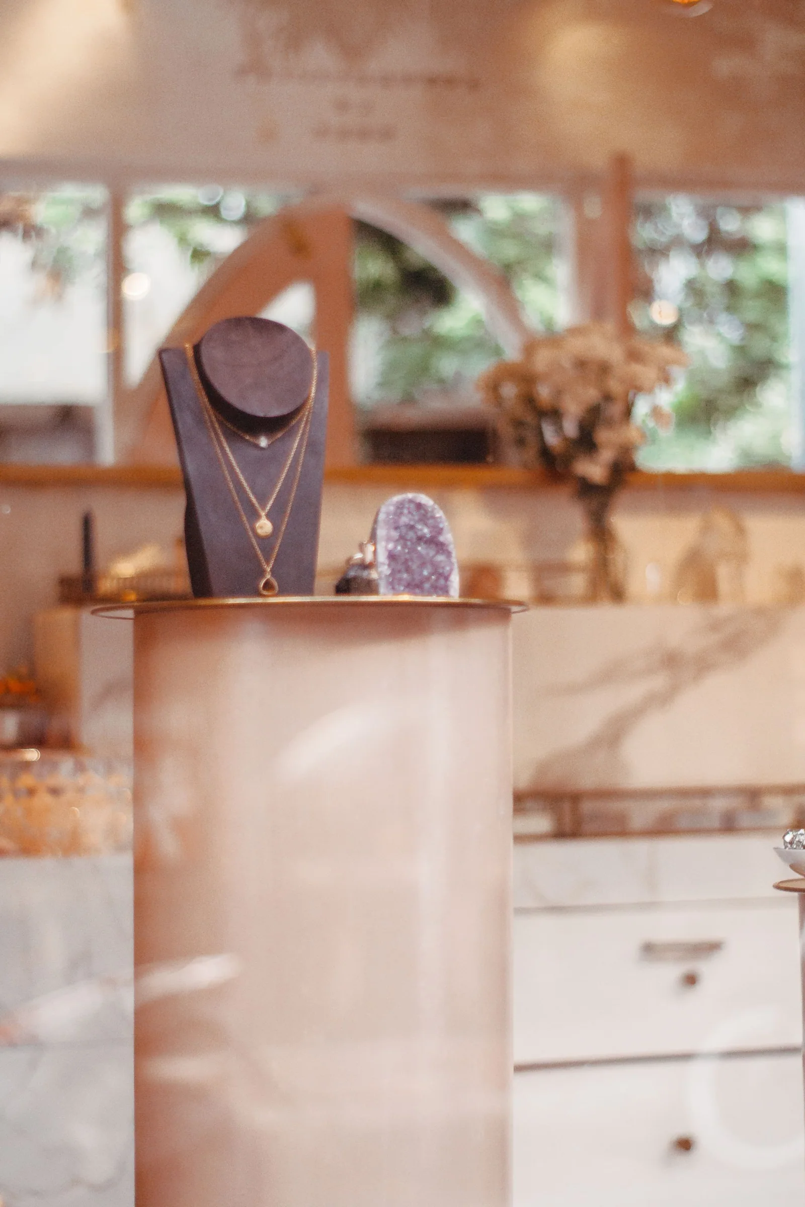 Layered necklaces on a dark display bust beside an amethyst specimen in a softly focused boutique interior.