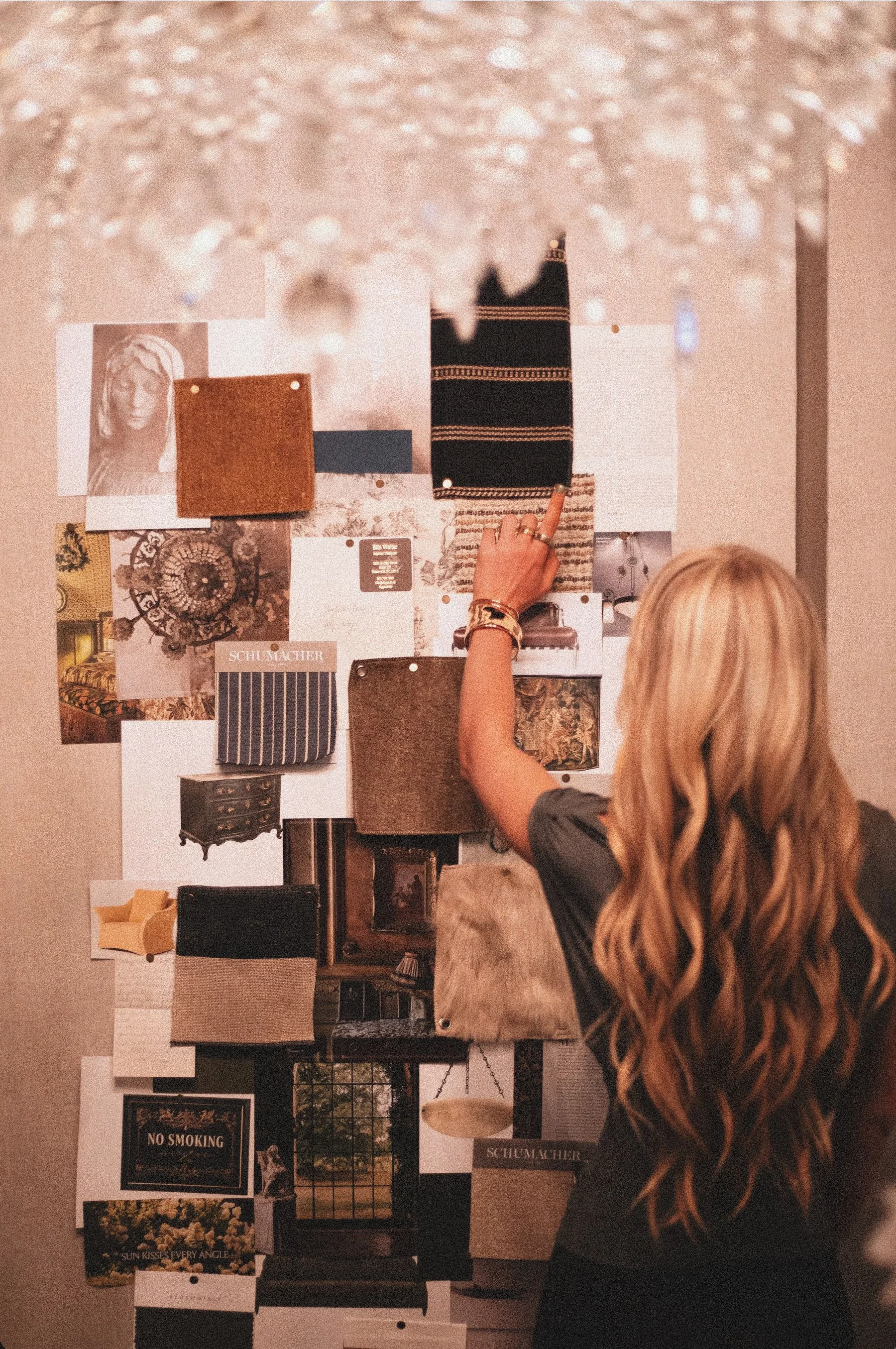 An interior designer pins textile swatches to a layered mood board beneath a glass chandelier.