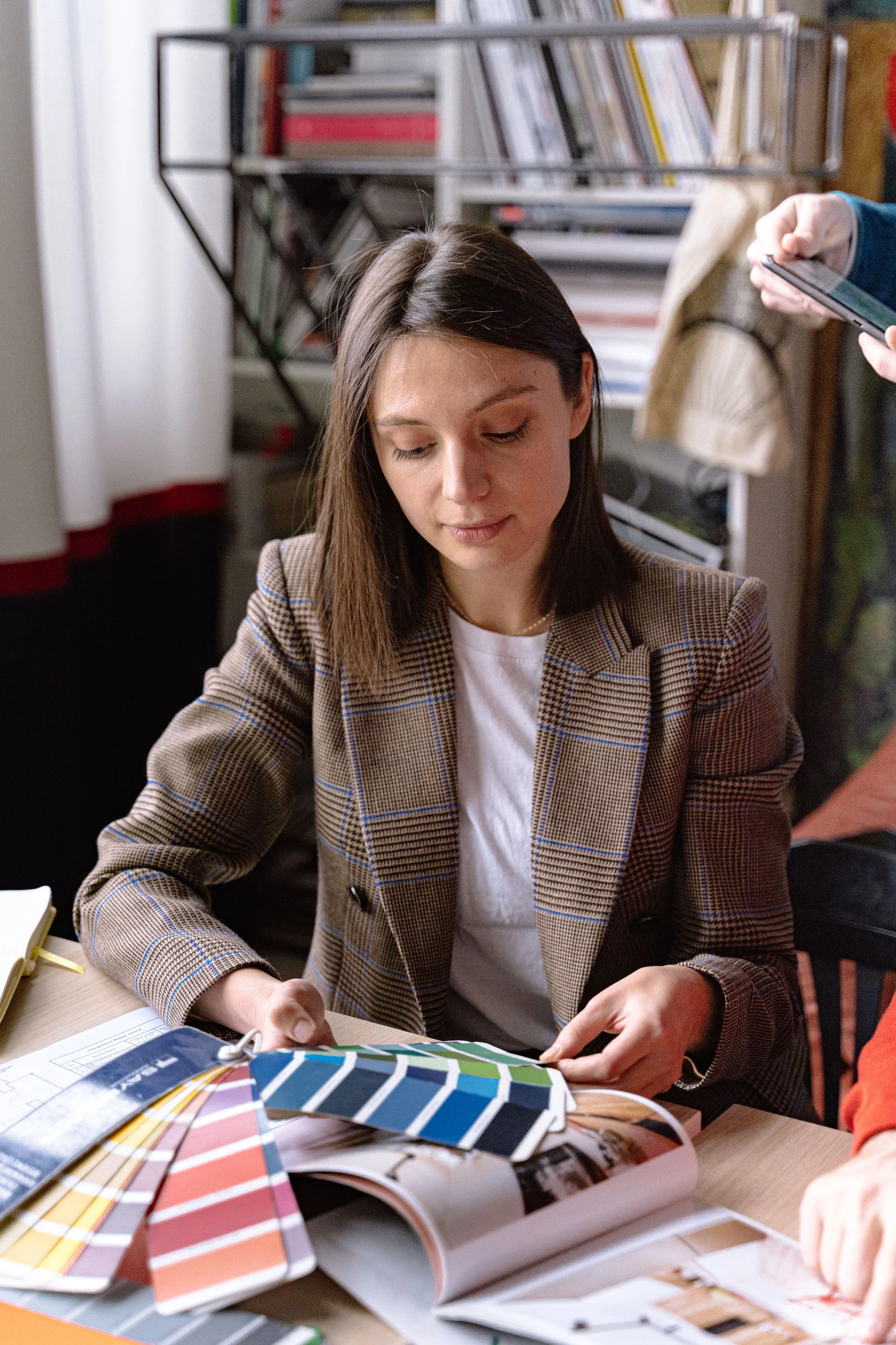 A woman in a tailored checked blazer compares blue and neutral paint samples at a studio table.
