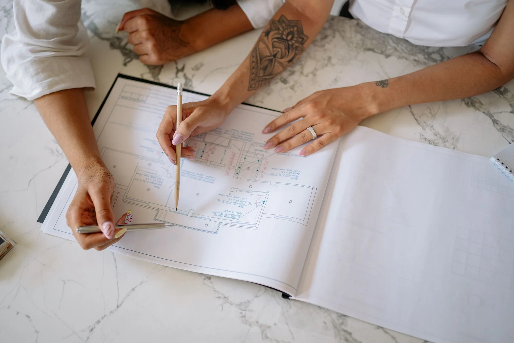 Two women use pencils to discuss an architectural floor plan on a marble table.