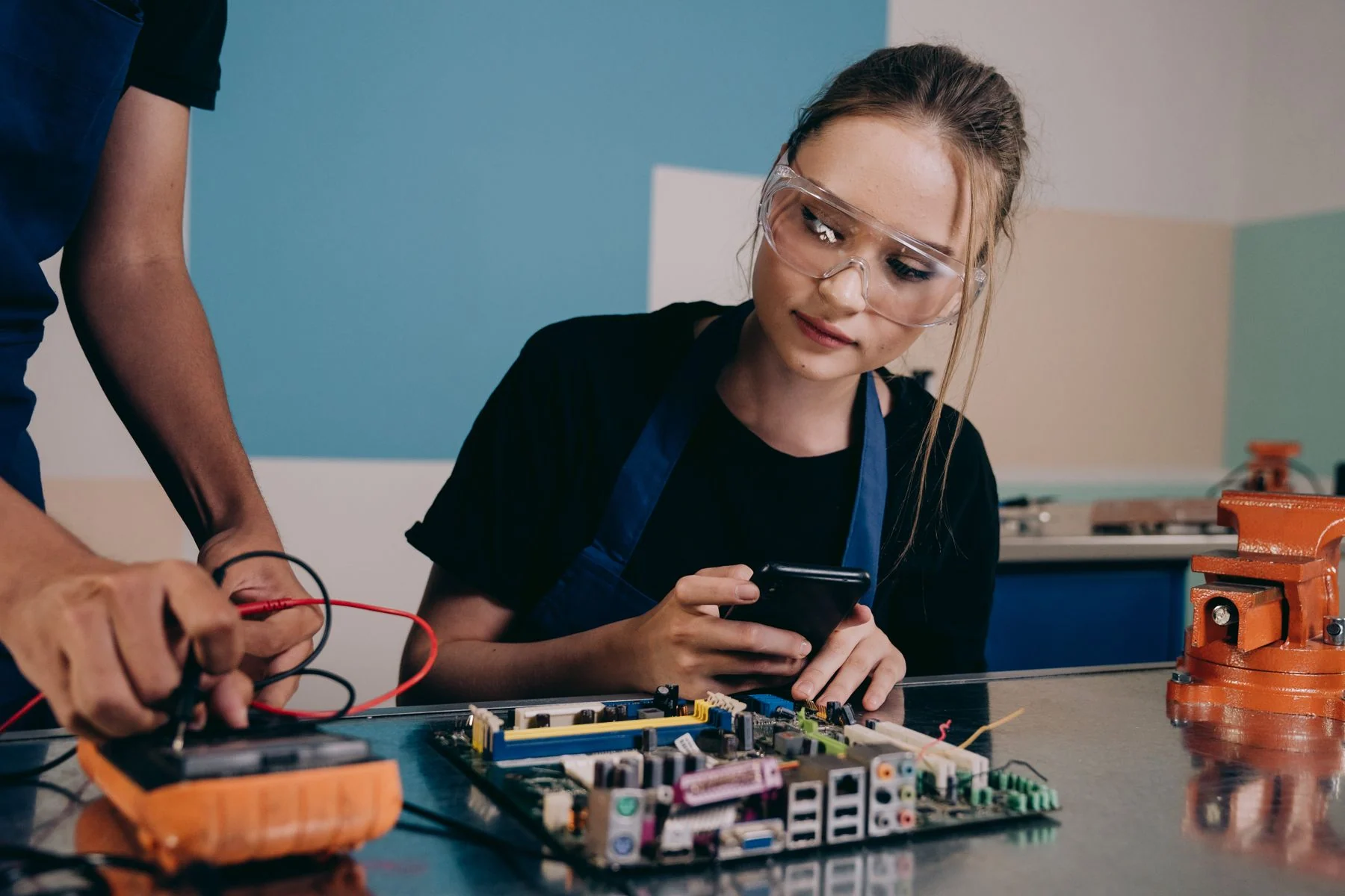A woman in safety glasses examines a circuit board at a workbench with a multimeter and a colleague.