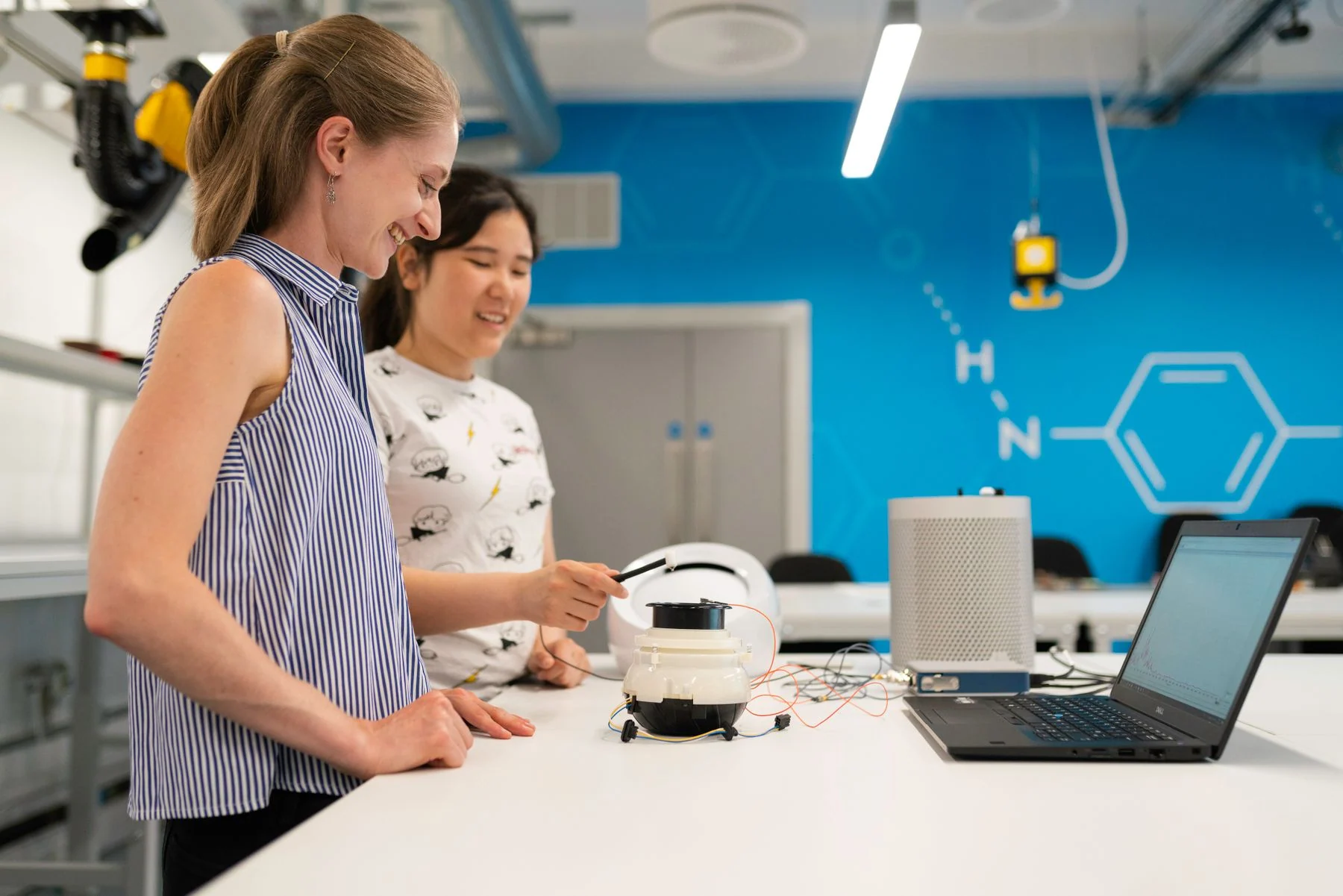 Two women investigate an electronic prototype and laptop at a bright laboratory bench.