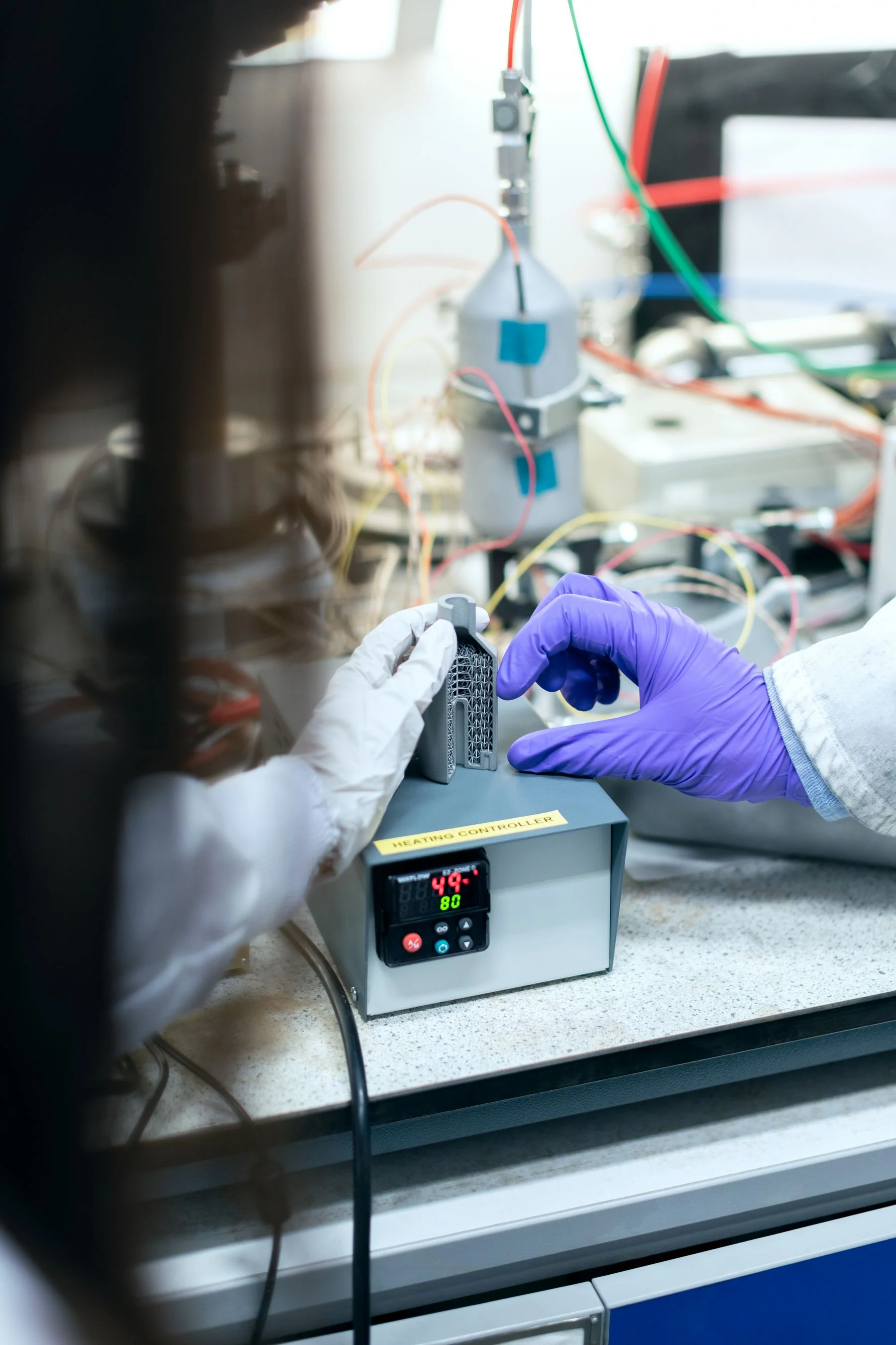 Gloved hands inspect a lattice sample beside a heating controller and laboratory equipment.