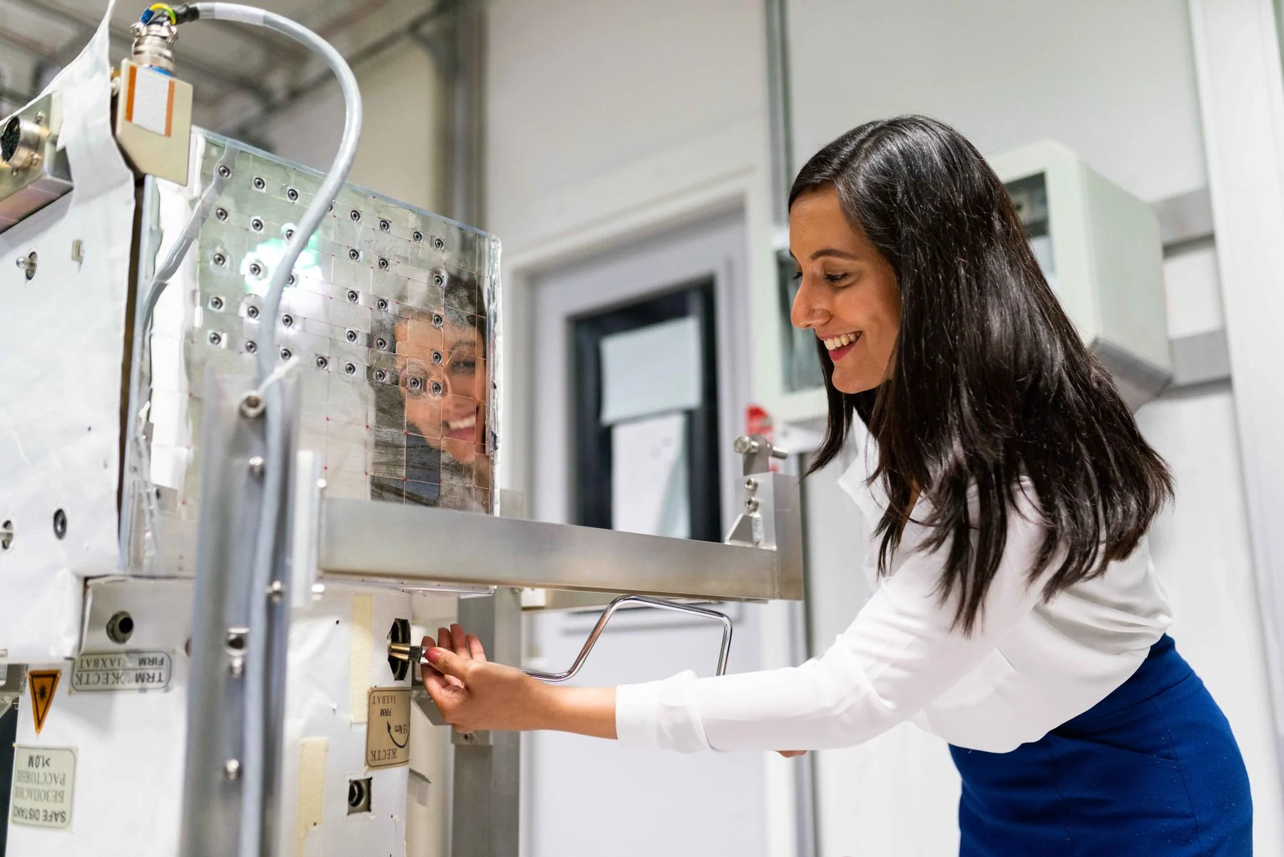 A smiling woman operates laboratory equipment beside a reflective metal enclosure.