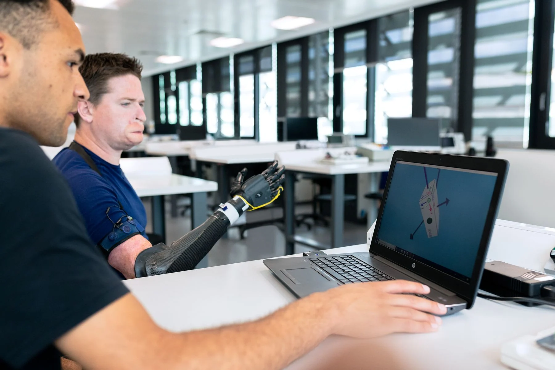 Two engineers review a prosthetic-arm design beside a laptop in a bright workspace.