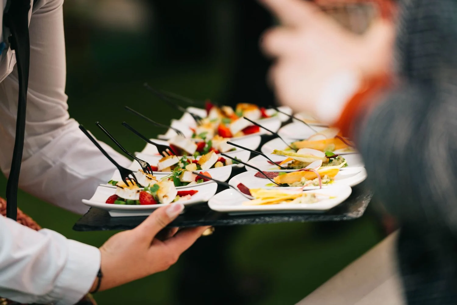 A server carries a tray of individually plated cheese, fruit and vegetable appetizers with small forks.