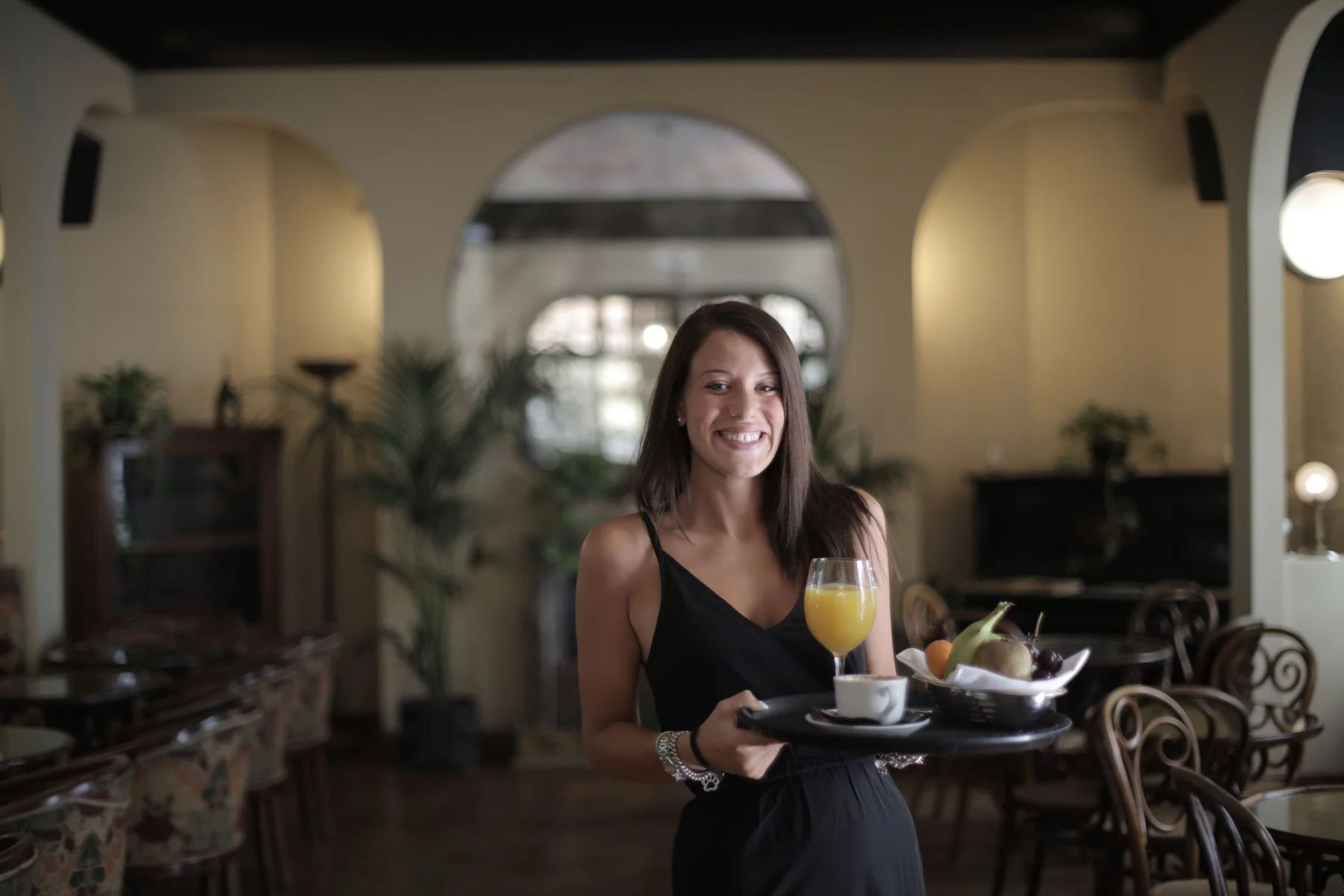 A smiling woman in a black dress presents a tray of fruit, juice and coffee in a restaurant.