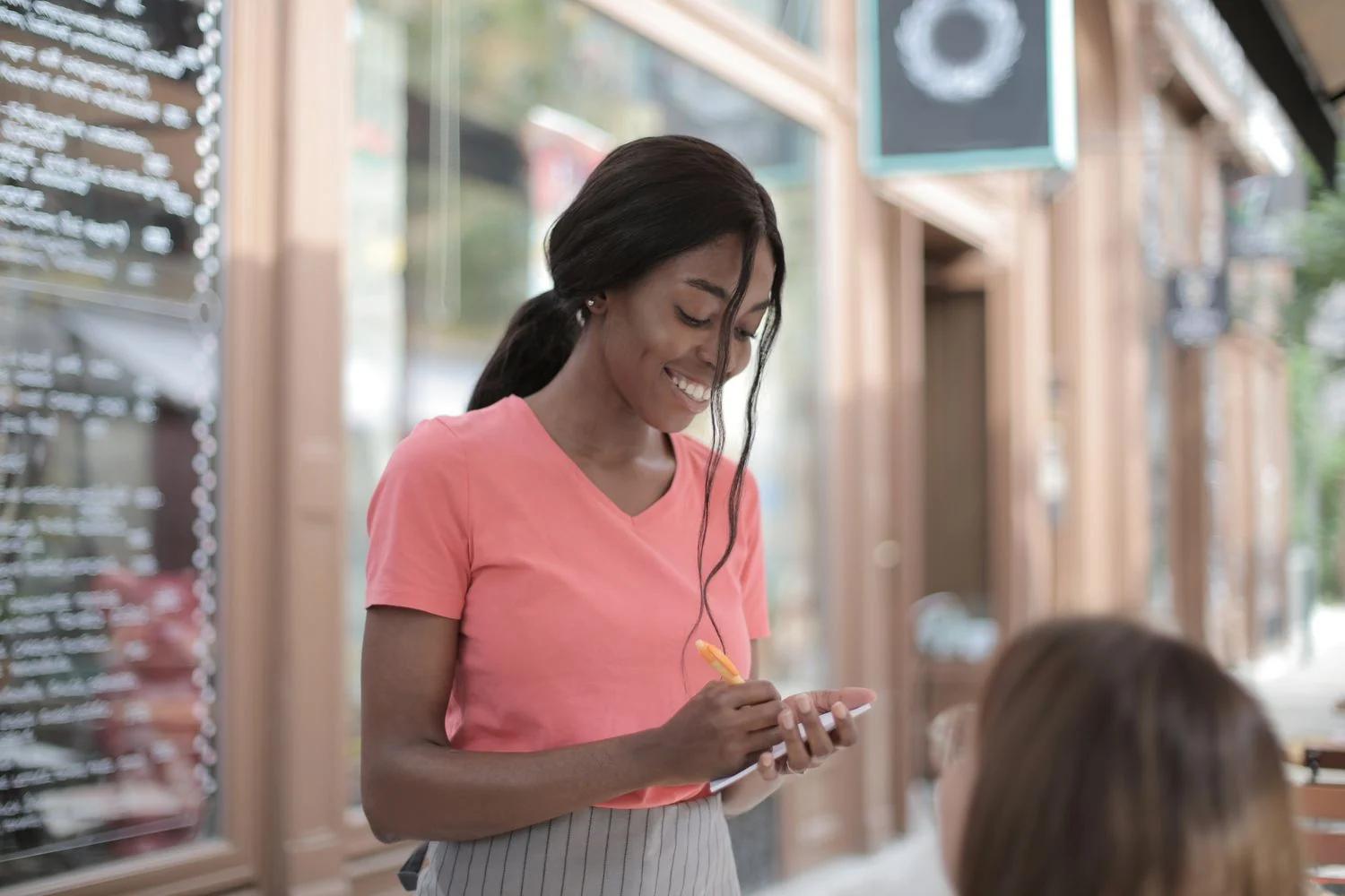 A smiling hospitality professional writes a customer's request in a notepad at an outdoor cafe.