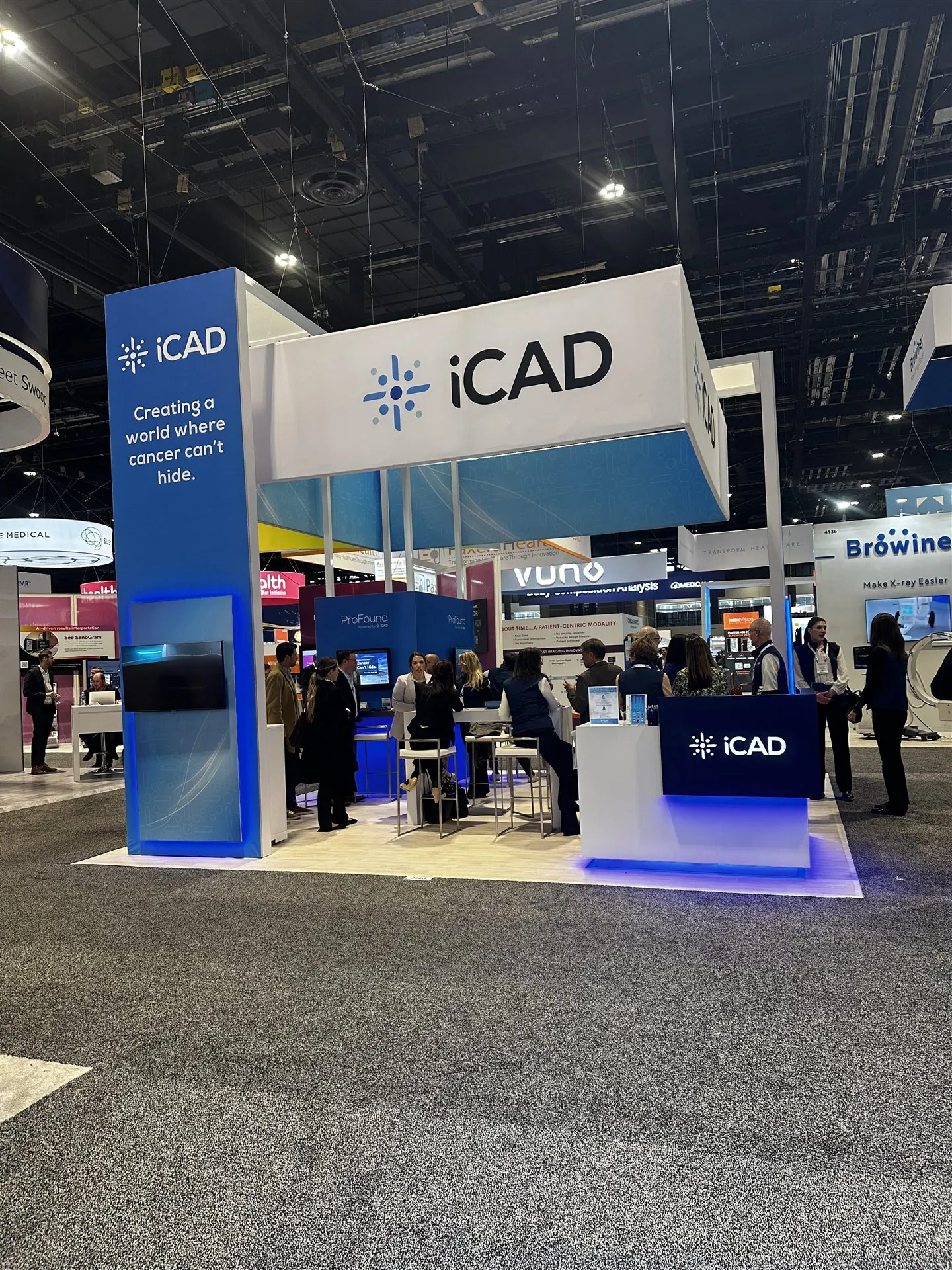 iCAD exhibit with a suspended square sign, blue cancer-detection graphics, an illuminated reception counter and visitors.