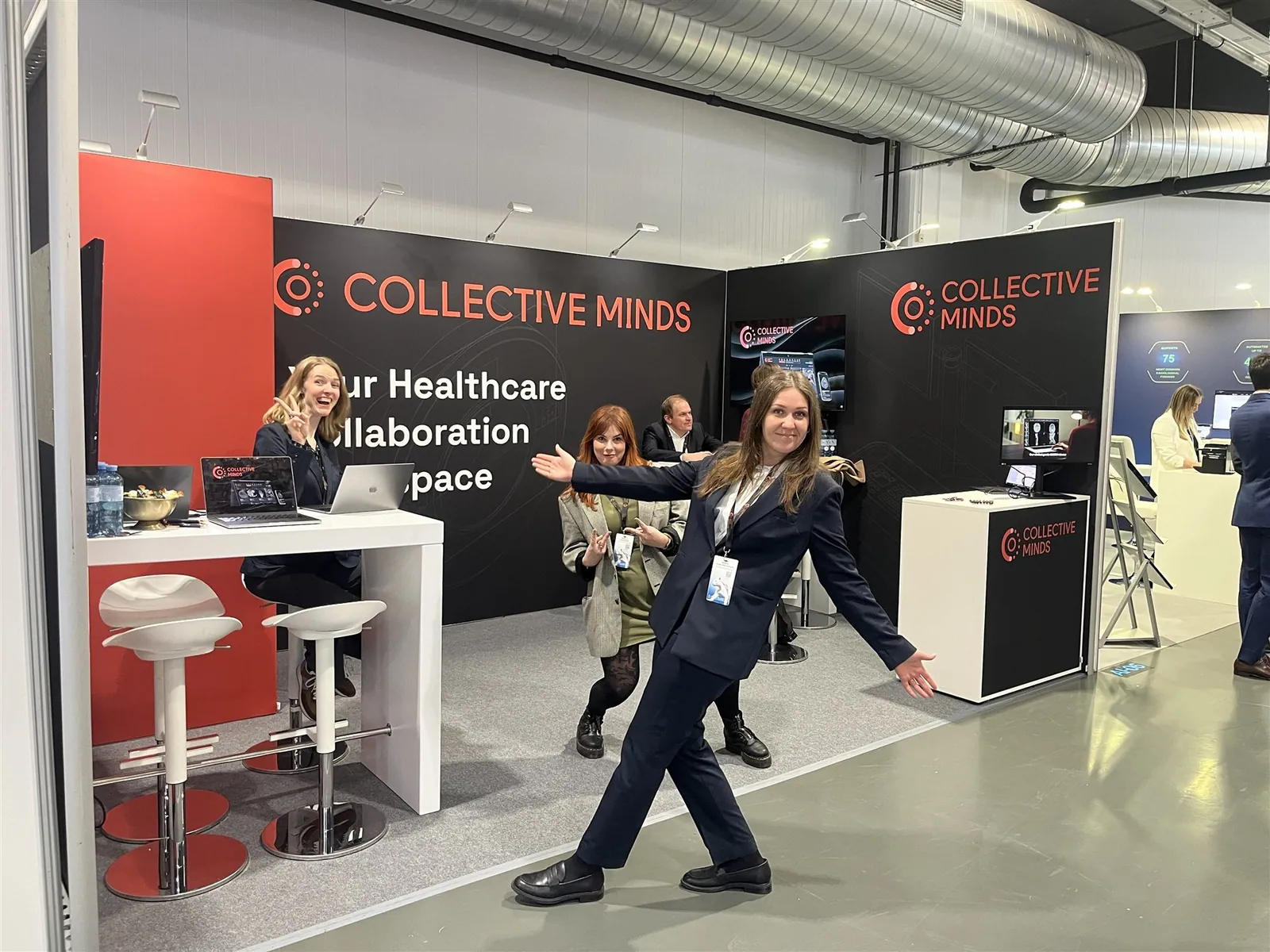 People posing inside Collective Minds' black-and-red healthcare collaboration exhibit beside laptops, screens and counters.